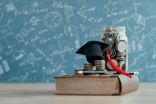 Clear jar full of quarters, along with piled quarters, mini graduation cap, and pencil stacked on top of a book laying on a tabletop in front of a chalkboard with formulas and figures written on it