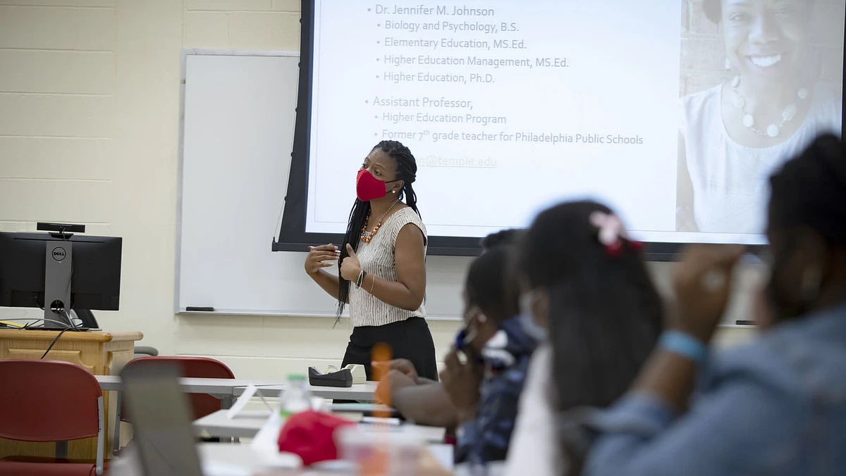 A woman teaching at the front of a classroom, wearing a red mask