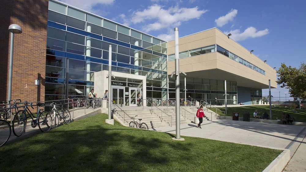 A modern building with large glass windows and numerous bicycles parked outside, with a few people walking or sitting nearby.