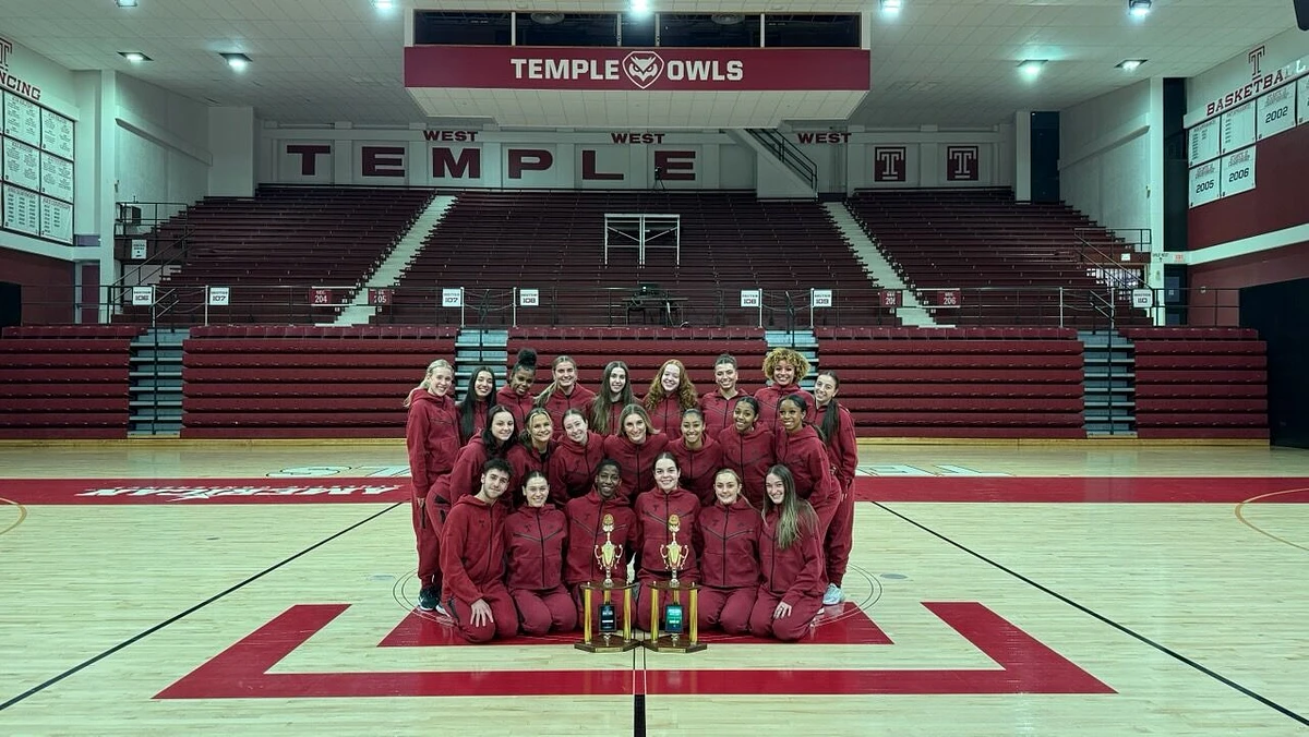 A group of people in red athletic outfits pose on a basketball court with three trophies at Temple University's gymnasium.