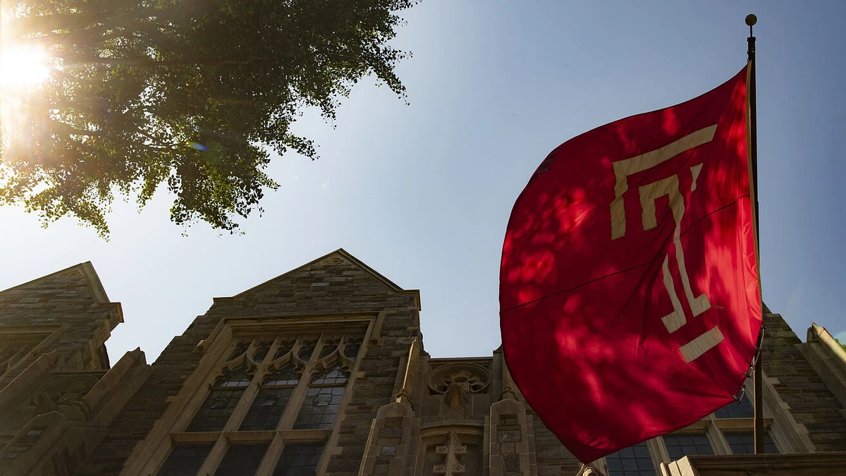 A red flag flutters in the sunlight next to a historic stone building with gothic-style windows.