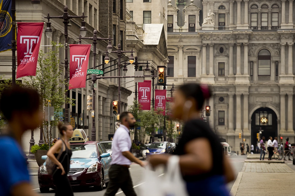 People are walking on a busy city street lined with Temple University banners and classic architectural buildings.