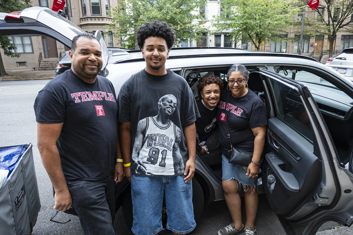 A family wearing Temple University shirts poses for a photo beside a car on a residential street.